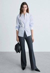 Camicia azzurro chiaro con colletto a punta, taschino sul lato sinistro e maniche arrotolate, abbinata a pantaloni svasati grigio scuro e una borsa nera.
