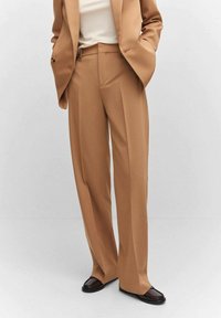 Pantalon beige ajusté avec une coupe droite, taille haute et léger pli devant, fabriqué dans un tissu lisse. Associé à un haut blanc.
