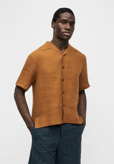 Άνδρας που φοράει καφέ κοντομάνικο πουκάμισο με κουμπιά και σκούρο μπλε παντελόνι με σχέδια, με τα χέρια στις τσέπες μπροστά σε απλό φόντο.