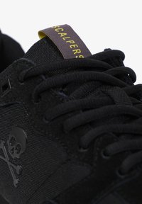 Zwarte sneaker met een gestructureerde bovenkant van stof, doodskoplogo, platte zwarte veters en een contrasterend label in geel en donkerpaars.