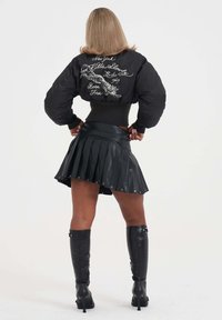 Giacca bomber nera con tigre ricamata e scritta sul retro, abbinata a una gonna in pelle nera plissettata e stivali neri alti fino alla coscia.