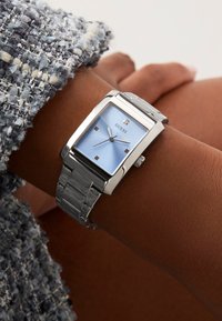 Montre rectangulaire argentée avec un cadran bleu clair, dotée de quatre marqueurs d'heure carrés et d'un bracelet en maillons métalliques. Portée au poignet.