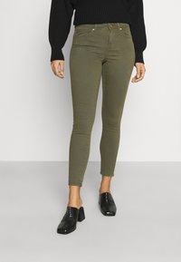 Jeans skinny verde-azeitona com uma textura suave, apresentando cinco bolsos, passantes de cinto e detalhes de costura subtis, combinados com mules pretos de salto.