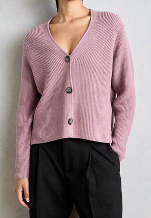 Kobieta ubrana w różowy, prążkowany kardigan z długim rękawem i trzema czarnymi guzikami, zestawiony z czarnymi, wysokim stanem spodniami.