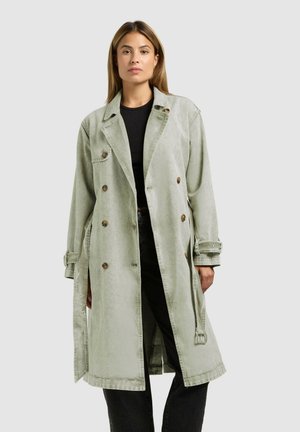 Žena v svetlozelenej dvojradovej trenčkovej bunde cez čierne tričko a čierne nohavice, stojaca pred jednoduchým sivým pozadím.