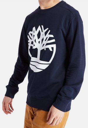 Mężczyzna ubrany w granatową bluzę z kapturem z dużym białym logo drzewa oraz brązowe spodnie, z rękami swobodnie opuszczonymi wzdłuż ciała, na jednolitym białym tle.