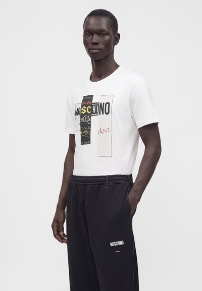 Άνδρας που φοράει ένα λευκό γραφιστικό t-shirt Moschino και μαύρο παντελόνι Moschino με το ένα χέρι στην τσέπη, στέκεται μπροστά σε απλό φόντο.