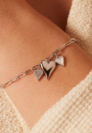 Silver chain bracelet with three heart-shaped charms resting on a wrist with light brown skin and beige knit fabric nearby.