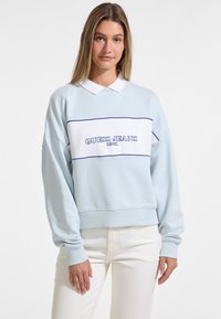 Vaalean sininen pitkähiha pulloveri, jossa on valkoiset ja tummansiniset raidat, esittelee "GUESS JEANS 1981" -logon, ribatuit hihansuut ja klassisen kauluksen.
