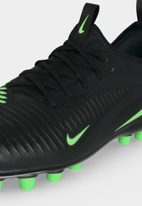 Μαύρο ποδοσφαιρικό παπούτσι Nike με πράσινες τάπες και πράσινα λογότυπα Nike swoosh στο πλάι και στη γλώσσα, που δείχνει κορδόνια και υφή στην επάνω πλευρά.