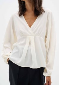 Blusa bianca con maniche lunghe a sbuffo e scollatura a V profonda. Il tessuto è liscio con una leggera lucentezza, presenta una vita arricciata e due pieghe.