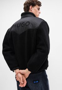 Čierna fleecová bunda s textúrovaným povrchom, obsahujúca lesklý čierny panel a vyšívané logo "HUGO" na zadnej strane. Viditeľné tmavé džínsy.