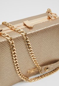 Borsa a mano rettangolare in metallo dorato con finitura texturizzata. Presenta una tracolla in catena dorata e una chiusura con gancio in metallo. Dettaglio del logo visibile.