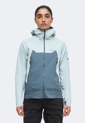 Femme portant une veste zippée bleu clair et grise avec un col haut, debout devant un fond uni, faisant face à l'avant.