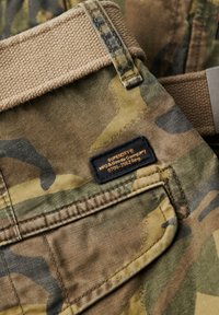 Pantalones cargo de camuflaje con cinturilla beige, con una textura tejida, detalles de etiqueta y múltiples bolsillos en un tejido de algodón.