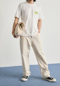 Valkoinen puuvillateepaita, jossa on pieni värikäs graafinen kuva rinnassa, yhdistettynä vaaleisiin beigeihin löysiä housuihin ja mustiin lenkkareihin.