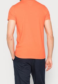 T-shirt arancione a maniche corte con una silhouette aderente, texture liscia e scollo rotondo. Indossato con pantaloni scuri, vista dal retro.