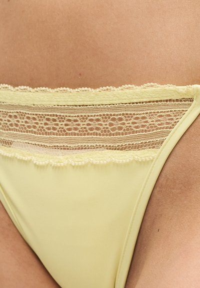 Κίτρινο εσώρουχο με δαντέλα στο ισχίο και το κάτω μέρος της κοιλιάς ενός ατόμου, που δείχνει περίπλοκη λουλουδένια δαντέλα στη μέση.