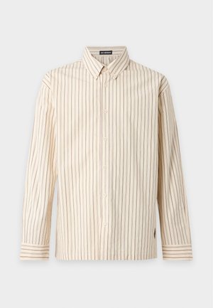 Jeune homme portant une chemise beige rayée à boutons, un pantalon cargo marron, une casquette marron et des chaussures noires, debout devant un fond uni.
