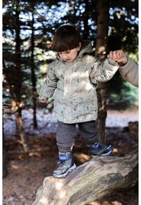 Jasnozielona puchowa kurtka w kwiaty, szare spodnie i ciemnoniebieskie buty. Dziecko stoi na pniu drzewa, trzymając się za rękę z dorosłym.