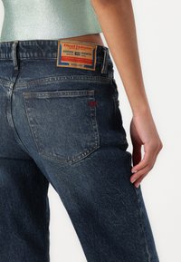 Primer plano de una persona que lleva unos jeans Diesel de color azul oscuro con un bolsillo trasero visible y una etiqueta de cuero de la marca en la cinturilla.