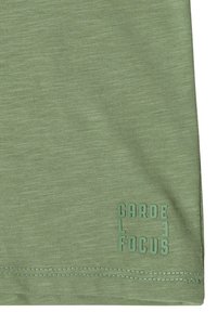 Tissu vert clair avec ourlet cousu et logo vert en relief portant l'inscription « GARDE FOCUS » en lettres majuscules près du coin inférieur droit.