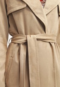 Beige lång kappa i mjukt mockakvalitet, med sjalkrage, bältad midja och sidofickor. Slät textur och avslappnad passform.
