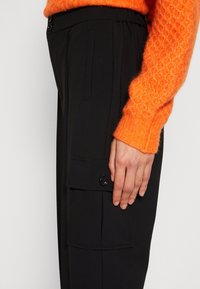 Pantalon cargo noir avec une poche latérale à fermeture par bouton, texture lisse, ceinture élastique et design discret.