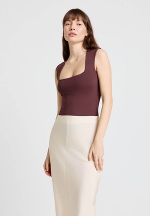 Donna con capelli castani che indossa un top bordeaux senza maniche con scollo quadrato e una gonna beige a vita alta su uno sfondo chiaro e uniforme.