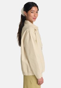 Licht beige jas met rits, gemaakt van zachte katoen. Heeft een kraag, elastische tailleband en twee zijzakken. Gladde textuur.