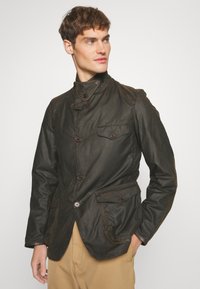Dark brown waxed jacket featuring a high collar, multiple buttons, two front pockets, and a sleek, fitted design. Matte texture.