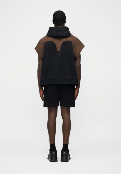 Model masculin văzut din spate purtând un hanorac cu glugă cu mâneci scurte, negru cu maro, pantaloni scurți negri, șosete negre și adidași negri, stând pe un fundal alb.