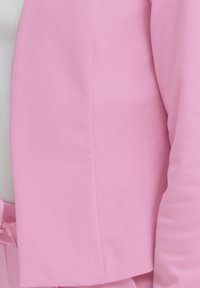 Persona valkā gaiši rozā žaketi virs baltas ribotas augšdaļas, tiek fokusēts uz torsu un roku zonu.