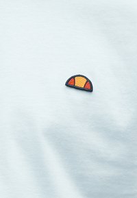 Ανοιχτό μπλε ύφασμα με ένα μικρό κεντημένο ημικυκλικό λογότυπο σε πορτοκαλί και κόκκινο με σκουρό μπλε περίγραμμα κοντά στο κέντρο.