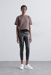 T-shirt castanho cropped com "EAT" em dourado, combinado com leggings pretas brilhantes e sapatilhas brancas. O modelo está posicionado contra um fundo cinza claro.