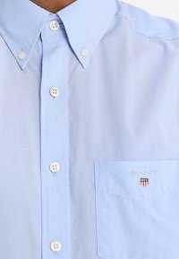 Camisa de botones azul claro con un bolsillo que presenta el logotipo y emblema bordados de "GANT". Textura de tela suave, diseño de cuello clásico y botones blancos.
