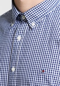 Camisa de cuadros vichy azul y blanca, con cuello abotonado, botones blancos y un pequeño logotipo en rojo y azul en el lado izquierdo del pecho.