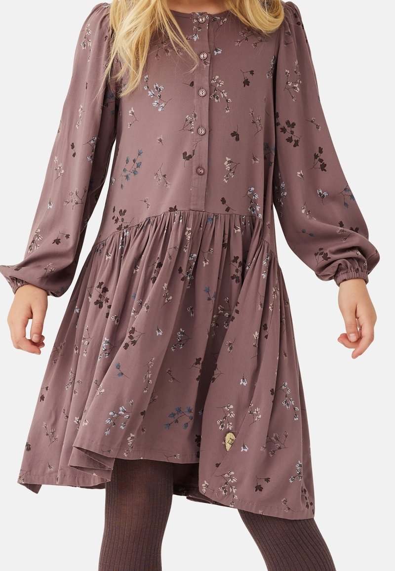 Fille portant une robe longue à manches longues mauve à fleurs avec des boutons sur la poitrine et une taille froncée, associée à des collants foncés côtelés.