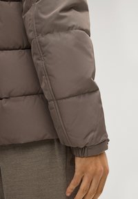 Bruine pufferjack met doorgestikte delen, ribbelmanchetten en een gladde afwerking. Zichtbare hand rustend op een grijze stoffen achtergrond.