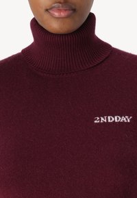 Osoba ubrana w bordowy sweter z prążkowanym golfem, z białym napisem "2ND DAY" na klatce piersiowej, na jednolitym tle.