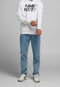 Felpa grigia con logo "Jack & Jones", abbinata a jeans in denim azzurro chiaro e sneakers bianche con accenti neri, in piedi su uno sfondo neutro.