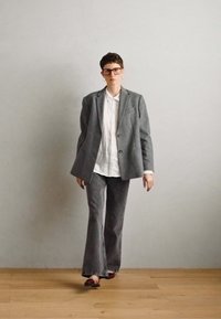 Blazer gri și pantaloni evazați asortați, realizați dintr-un material texturat, asociați cu o cămașă albă cu guler. Pantofii roșii cu talpă plată completează ținuta.