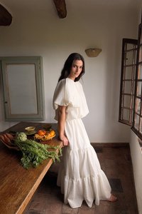 Abito bianco a strati, texturizzato, con maniche a sbuffo; indossato a piedi nudi. Verdura fresca su un tavolo di legno in un ambiente rustico interno.
