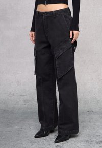 Pantalones cargo negros con piernas anchas, múltiples bolsillos, detalles de costura y un ajuste relajado. Fabricados en tela de mezclilla con un acabado desgastado.