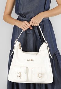 Bolso de mano de piel blanca con dos bolsillos frontales, herrajes dorados y cierre con cremallera. Textura suave y diseño holgado con asas cortas.