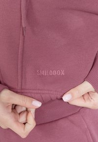 Felpa fucsia scuro con polsini e orlo a coste, con il logo "SMILODOX" in rilievo sul davanti, che mostra un primo piano del tessuto testurizzato.