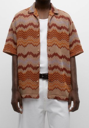 Chemise en maille à manches courtes avec des motifs zigzag marron, orange et beige portée sur un débardeur blanc et un pantalon blanc avec une ceinture noire.