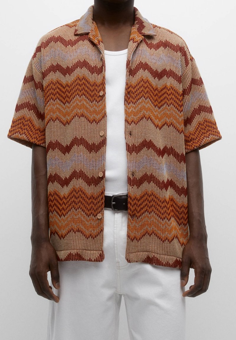 Chemise en maille à manches courtes avec des motifs zigzag marron, orange et beige portée sur un débardeur blanc et un pantalon blanc avec une ceinture noire.