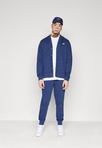 Σκούρο μπλε cardigan της Nike με κούμπωμα, πάνω από ένα λευκό μπλουζάκι, συνδυασμένο με αντίστοιχες σκούρες μπλε φόρμες και λευκά αθλητικά παπούτσια.