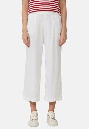 Vrouw draagt losse witte cropped broek, rood-wit gestreept shirt en gebroken witte sneakers, staand tegen een effen achtergrond.